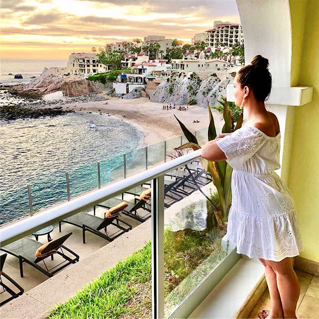 A brunette woman in a white dress on a balcony looking out at the ocean at Sirena del Mar by Welk Resorts in Cabo San Lucas
